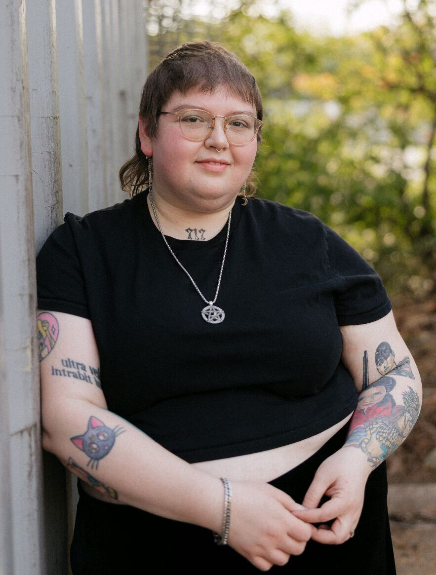 lydia-henderson-ma-atr-p-art-therapist-nashville-tn Lydia Henderson, MA, ATR-P, art therapist in Nashville, Tennessee, standing outdoors beside a light-colored wall with greenery in the background.