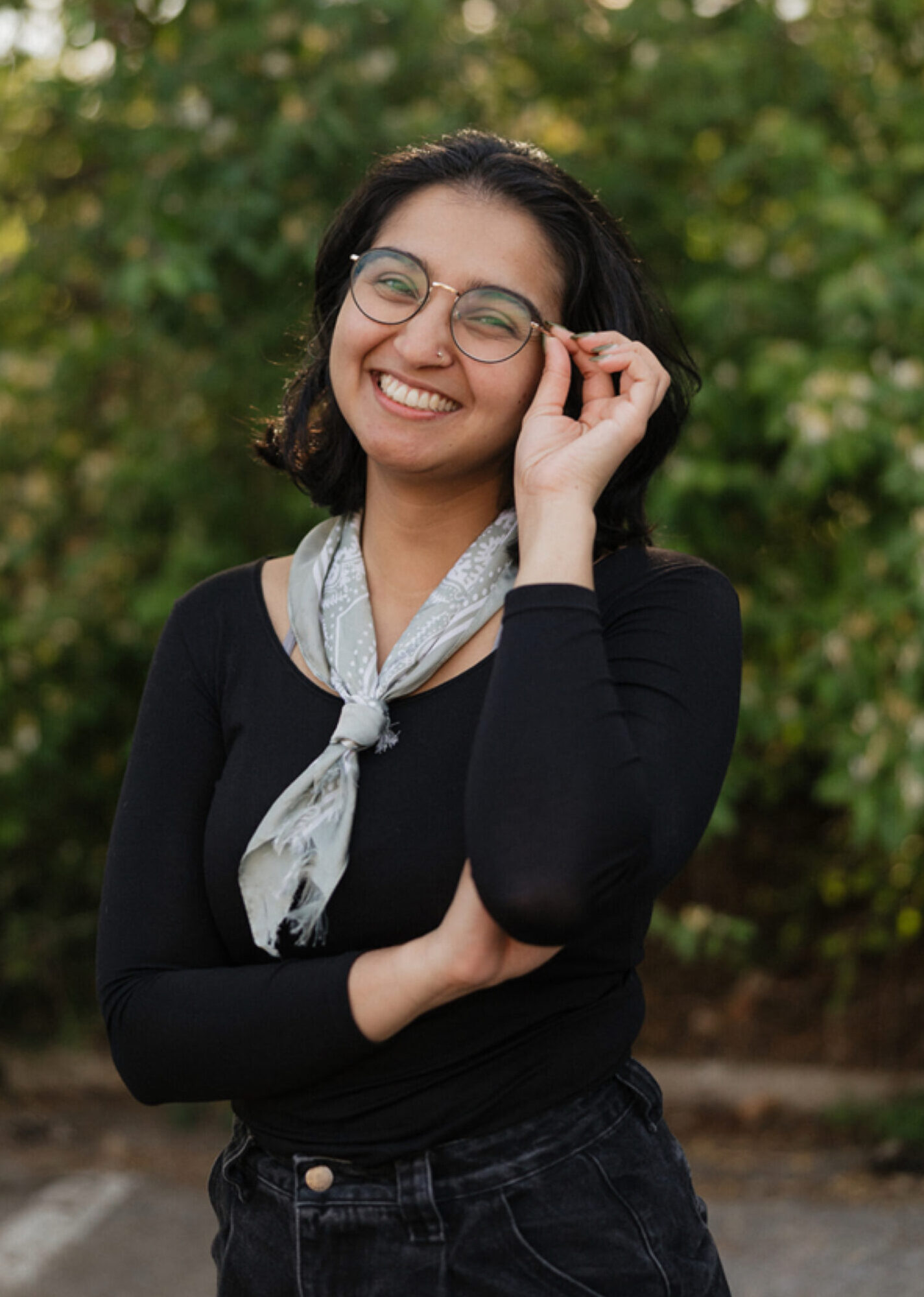 summer-sandhu-therapist-intern-nashville-tn Summer Sandhu, therapist intern in Nashville, Tennessee, smiling outdoors and wearing glasses, with greenery in the background.